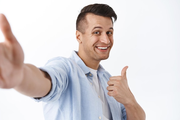 Portrait of handsome friendly guy show thumbup as taking selfie in front of white background smiling broadly give recommendation of good place stream live blog videocalling friends