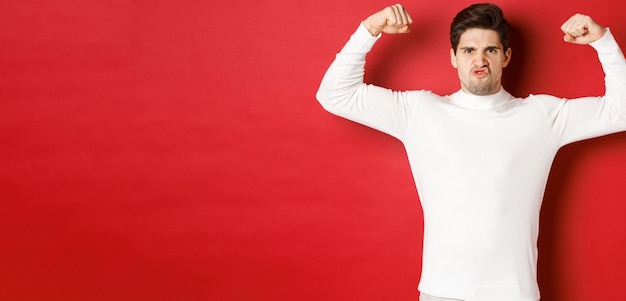 Portrait of handsome and funny guy in white sweater, flex biceps and looking encouraged, showing strong muscles, standing over red background
