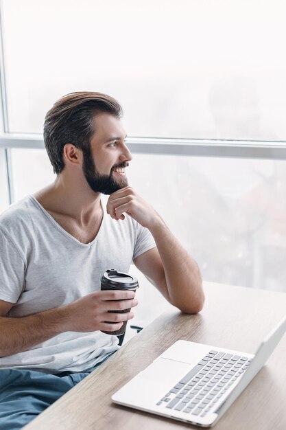 Portrait of handsome happy bearded young businessman in grey T-shirt, sitting in office and have a break with cup of coffee. He uses his laptop, looking out the window with smile