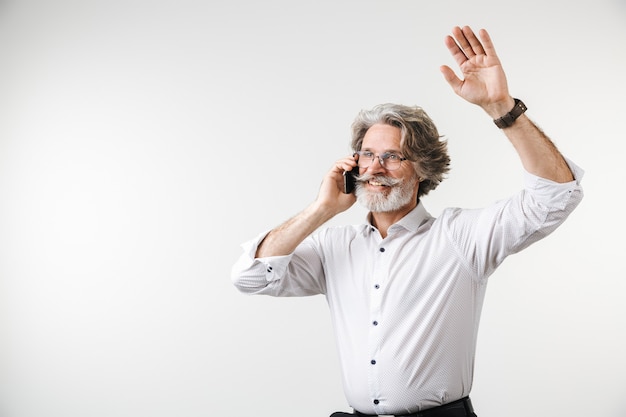 Ritratto di un bell'uomo d'affari maturo felice vestito con abiti formali in piedi isolato su un muro bianco, agitando la mano mentre parla al telefono cellulare