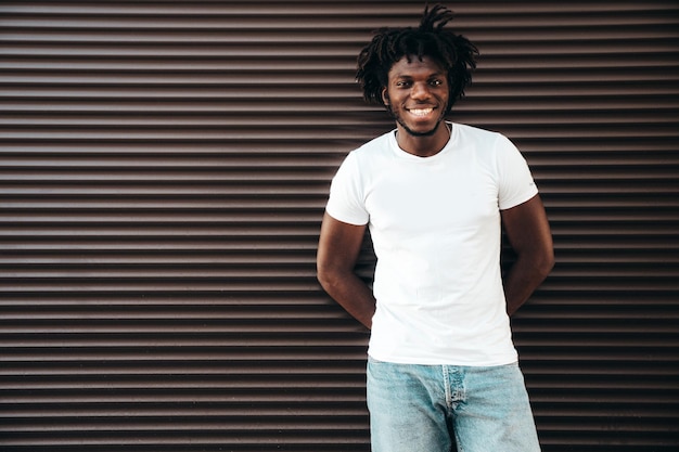 Portrait of handsome hipster modelunshaven african man dressed in white summer tshirt and jeans fashion male with dreadlocks hairstyle posing near roller shutter wall in the street