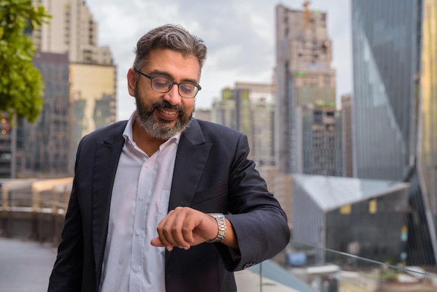 Portrait of handsome Indian businessman in city at rooftop garden checking time from wristwatch