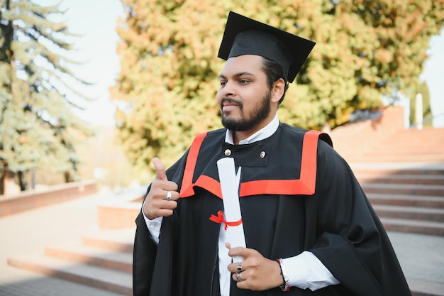 Portrait of handsome indian graduate student in graduation glow with diploma