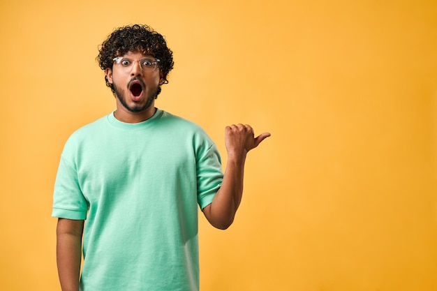 Portrait of a handsome Indian man with curly hair in a turquoise tshirt and glasses standing on a yellow background and pointing to the side Emotion of shock surprise