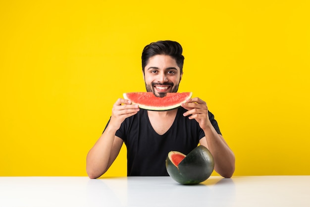 Portrait of a handsome Indian young man smiling and eating fresh watermelon or tarbooj while sitting at table or isolated over wooden floor