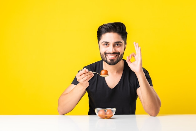 Portrait of a handsome Indian young manÃÂ eating sweet Gulab Jamun against yellow background