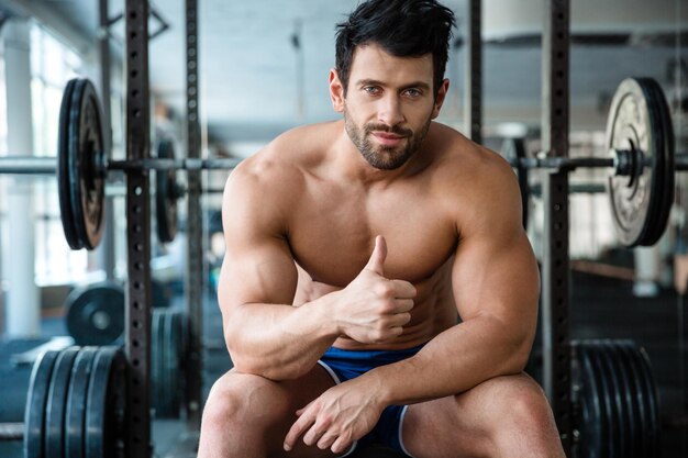 Portrait of a handsome male bodybuilder sitting on the bench in fitness gym and showing thumb up