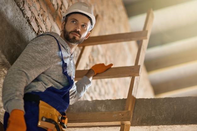 Photo portrait of handsome male builder in blue overalls and hard hat looking away climbing down