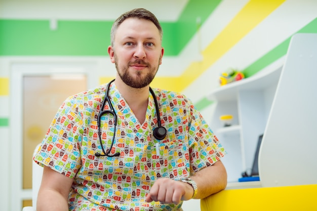 Portrait a handsome male doctor with stethoscope sitting in childish room. Pediatrician with stethoscope looking at camera. Close-up. Healthcare and medicine.