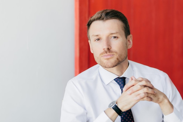 Portrait of handsome male employee has stubble holds hands together looks thoughtfully at camera contemplates about business project poses against red and white background with copy space