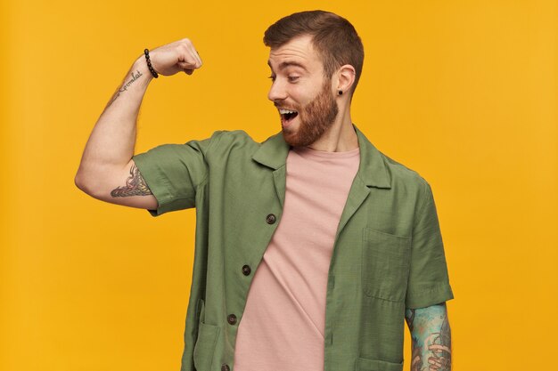 Portrait of handsome male with brunette hair and bristle. wearing green short sleeves jacket. has tattoos. showing his biceps and watching at it excited. stand isolated over yellow wall