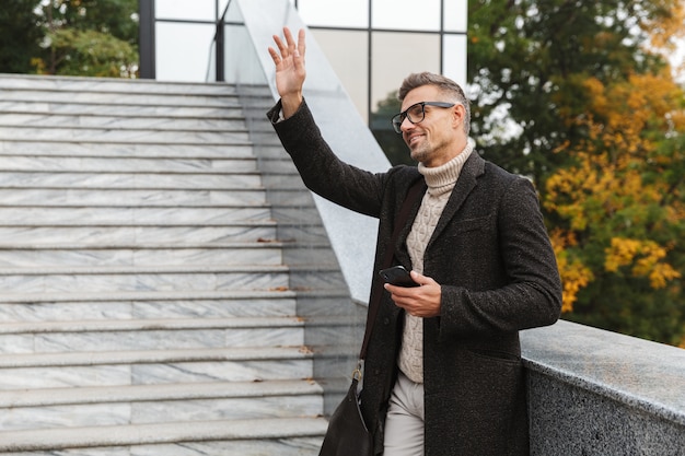 Portrait of handsome man 30s wearing eyeglasses, walking through city street and using cell phone