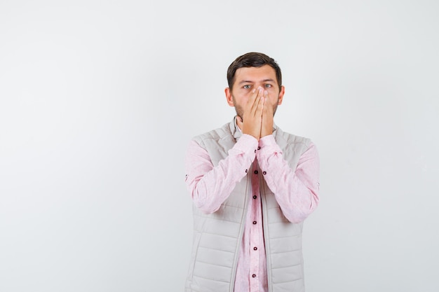 Portrait of handsome man keeping hands on mouth and nose in vest, shirt and looking shocked 