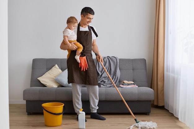 Portrait of handsome man wearing casual attire and brown apron cleaning house with mop and holding toddler daughter in hands doing housework washing floor