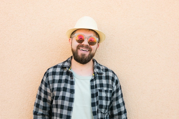 Ritratto uomo bello che indossa cappello estivo e occhiali da sole e camicia a quadri sorridente felice vicino al muro trav