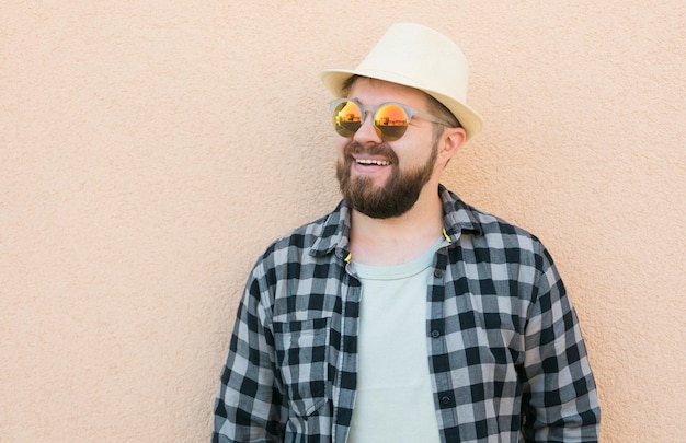 Ritratto di bell'uomo che indossa cappello estivo e occhiali da sole e camicia a quadri sorridente felice vicino al muro vacanze di viaggio e concetto di vacanza estiva