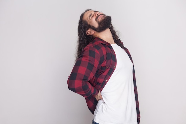 Portrait of handsome man with beard and black long curly hair in casual style, checkered red shirt standing holding his painful spine and feel backache. indoor studio shot, isolated on grey background
