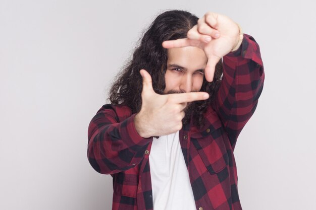 Ritratto di bell'uomo con barba e capelli ricci lunghi neri in stile casual, camicia rossa a scacchi in piedi guardando e facendo la cornice della fotocamera con le mani. foto in studio al coperto, isolata su sfondo grigio