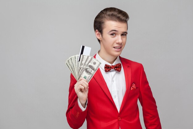 Portrait of handsome man with stylish hairdo in red jacket and\
bow tie showing bunch of dollars and bank card looking with\
confused doubtful expression studio shot isolated on gray\
background