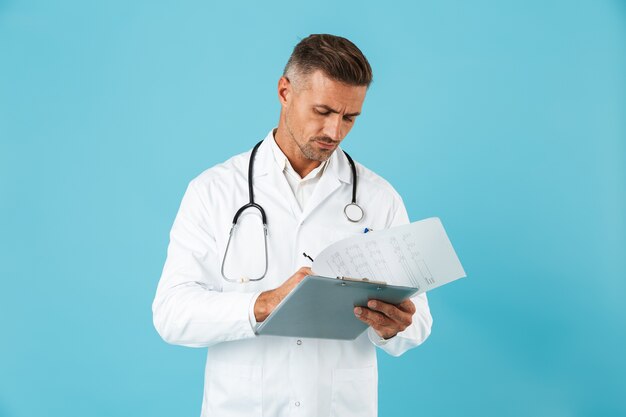 Portrait of handsome medical doctor with stethoscope holding health card, standing isolated over blue wall