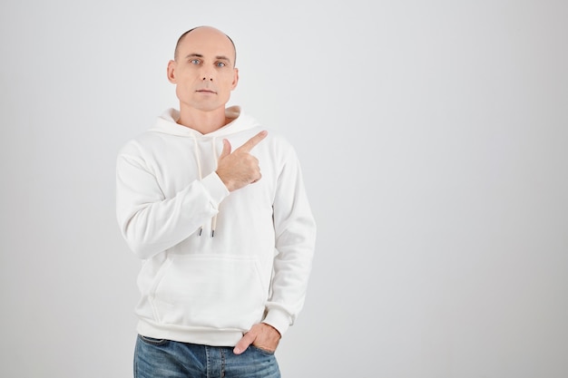 Portrait of handsome middle-aged an in white hoodie looking at camera and pointing away, isolated on white