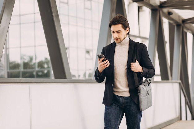 Portrait of handsome modern male businessman in suit walking smiling and reading or writing message on phone on background of urban buildings and offices