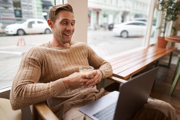 Portrait of handsome modern man sitting in coffee shop with laptop looking at screen with happy face