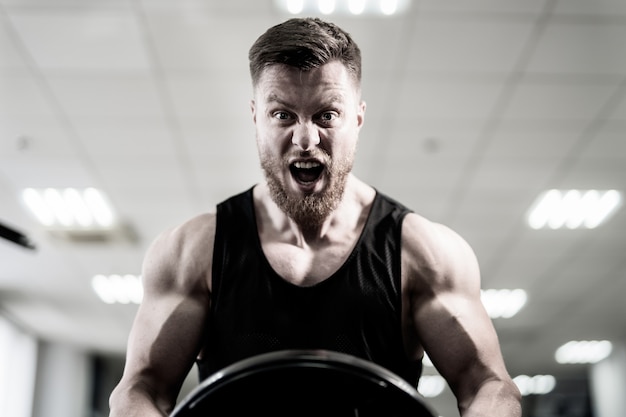 Ritratto del powerlifter muscolare bello con il disco del peso in sue mani nella palestra di sport. culturista con disco pesante allena i suoi bicipiti con la bocca aperta. avvicinamento. bianco e nero.