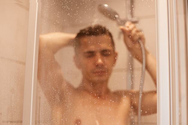 Portrait of handsome naked young man taking shower in bathroom, washing hair, keeping eyes closed, relaxing and enjoying while standing under hot water.