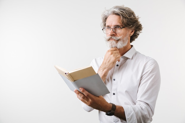 Portrait of a handsome pensive mature businessman dressed in formal wear standing isolated over white wall, reading a book