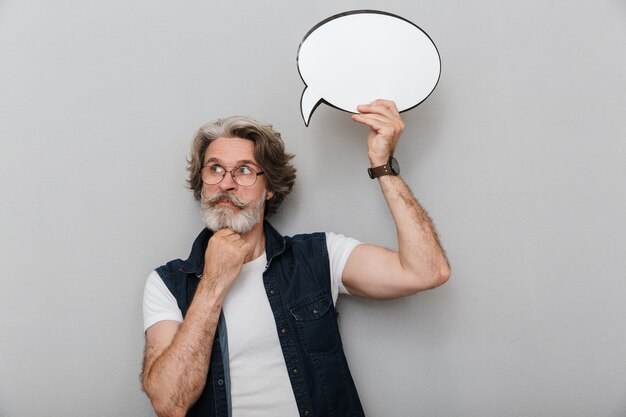 Portrait of a handsome pensive stylish mature man wearing a vest standing isolated over gray wall, holding empty speech bubble