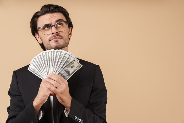 portrait of a handsome pensive young businessman wearing suit standing isolated over beige wall, showing money banknotes