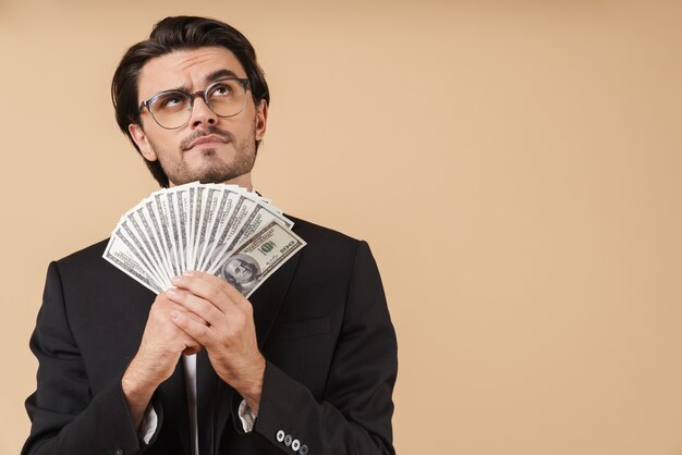 Portrait of a handsome pensive young businessman wearing suit standing isolated over beige wall, showing money banknotes