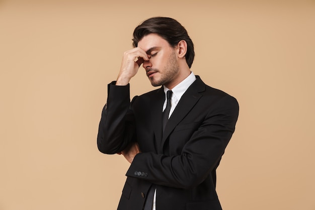 Portrait of a handsome pensive young businessman wearing suit standing isolated over beige wall, thinking