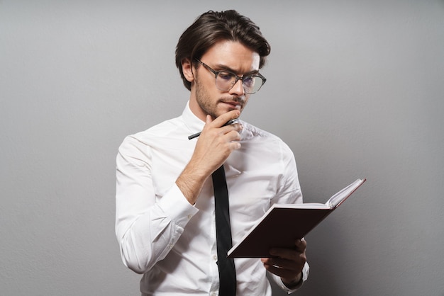 Portrait of a handsome pensive young businessman wearing suit standing isolated over gray wall, holding a journal