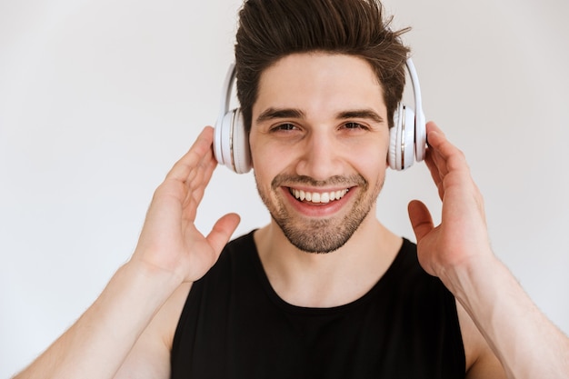 Ritratto di un giovane sportivo sorridente compiaciuto bello isolato sopra la musica d'ascolto della parete bianca con le cuffie.