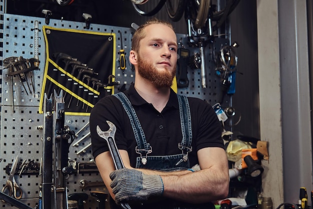 portrait of a handsome redhead male with beard and haircut wearing jeans coverall, holds steel wrench, standing in a workshop against a wall tools.