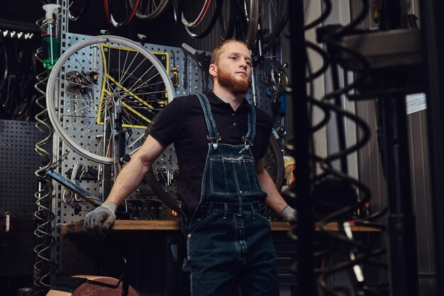 portrait of a handsome redhead male with beard and haircut wearing jeans coverall, standing near bicycle wheel in a workshop.