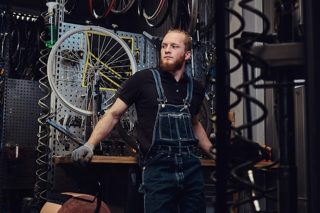 portrait of a handsome redhead male with beard and haircut wearing jeans coverall, standing near bicycle wheel in a workshop.