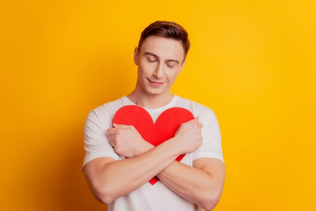 Ritratto di un bel ragazzo romantico ispirato e rilassato che abbraccia la figura del cuore su sfondo giallo