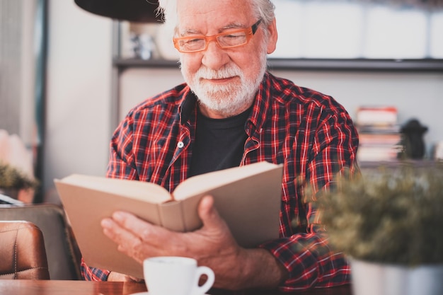 Portrait of handsome senior bearded man sitting at cafe table reading a book enjoying a coffee cup
