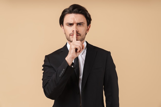 Portrait of a handsome serious young businessman wearing suit standing isolated over beige wall, showing peace gesture