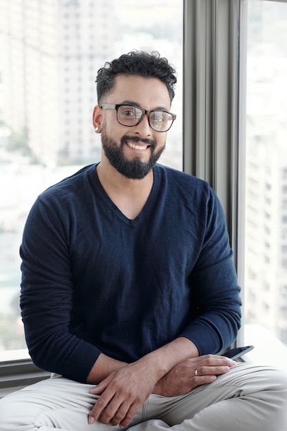 Portrait of handsome smiling man in glasses sitting on window sill and looking at camera