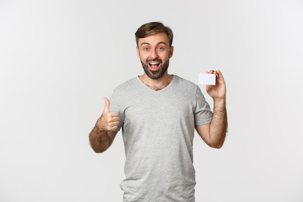 Портрет красивого улыбающегося человека в серой футболке, показывающего кредитную карту, поднимающего вверх палец в знак одобрения