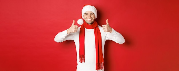 Портрет красивого улыбающегося мужчины в санта-шарфе и шляпе, показывающего большой палец вверх, празднует рождественский стенд ...