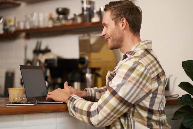 Portrait of handsome smiling man sitting with laptop in a cafe typing working remotely from