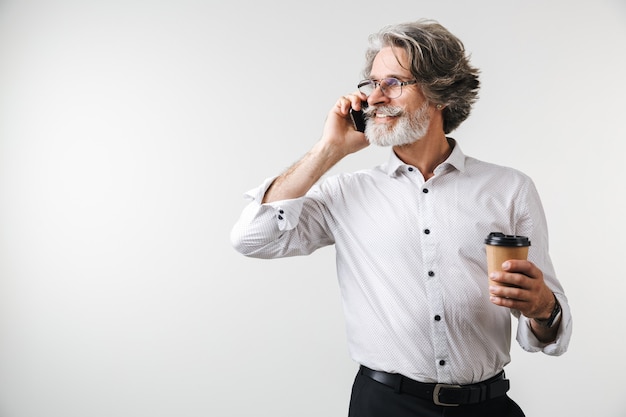 Ritratto di un bel uomo d'affari maturo sorridente vestito in abiti formali in piedi isolato su un muro bianco, parlando al telefono cellulare mentre beve caffè da asporto