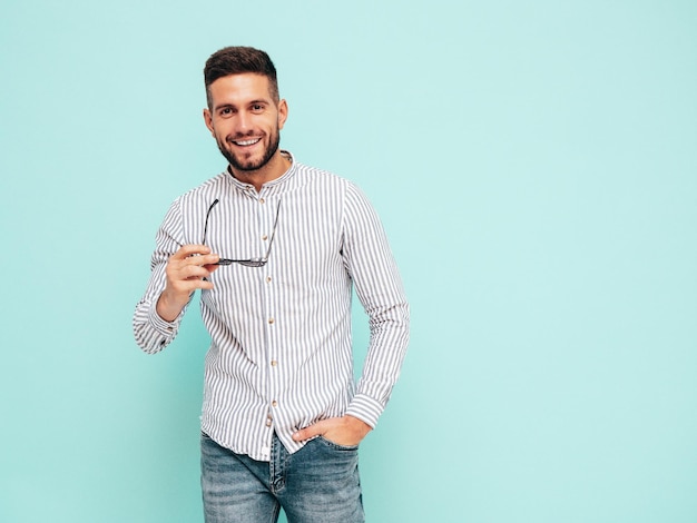 Portrait of handsome smiling model Sexy stylish man dressed in shirt and jeans Fashion hipster male posing near blue wall in studio Cheerful and happy