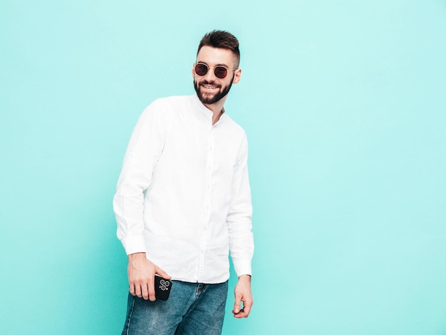 Ritratto di modello sorridente bello uomo alla moda sexy vestito con camicia bianca e jeans moda uomo hipster in posa vicino al muro blu in studio isolato in occhiali da sole