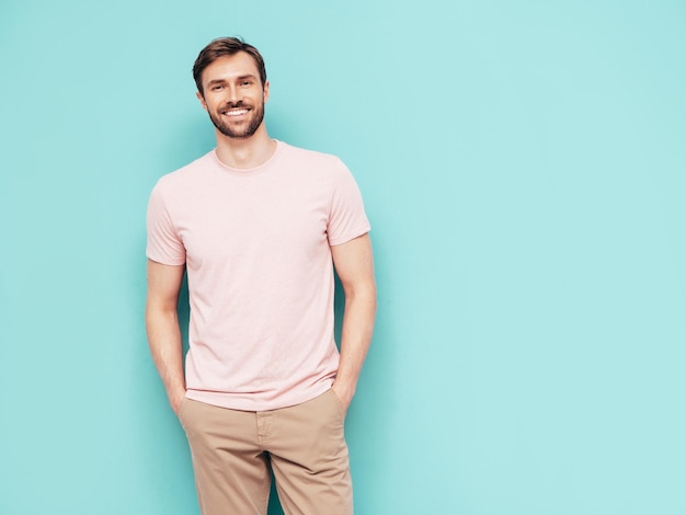 Portrait of handsome smiling stylish hipster lambersexual model Sexy man dressed in pink Tshirt and trousers Fashion male isolated on blue wall in studio